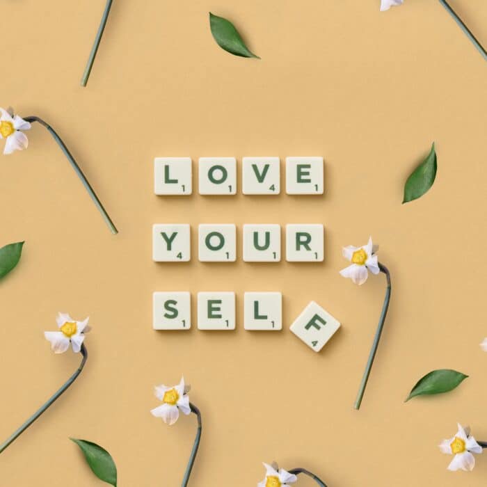Scrabble tiles spelling 'Love Yourself' surrounded by flowers and leaves on a flat surface.